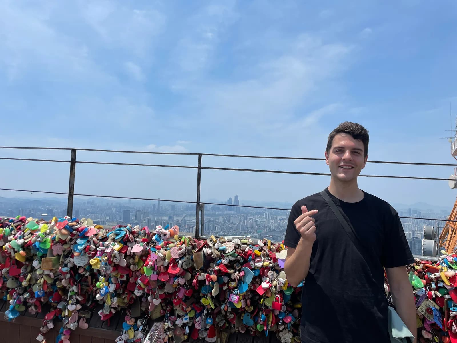 Nick making a finger heart beside a wall of love locks above Seoul